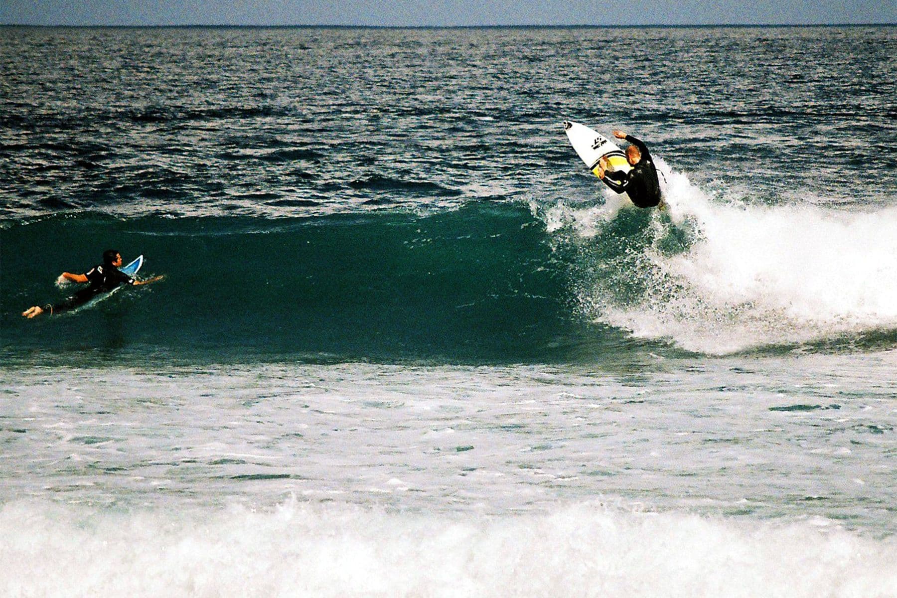 surfer doing a re-entry on a small right hand wave as another surfer paddles over the shoulder and watches